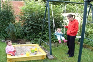 A child playing in a sandbox with toys, a swing set, and a garden in the background. - Holiday apartment in Suhl with swimming pool (Suhl)