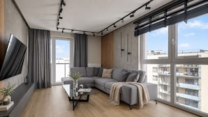 A modern living room with a large gray sectional sofa, a glass coffee table, and a large window offering a view of the cityscape.