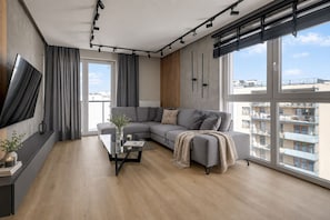 A modern living room with a large gray sectional sofa, a glass coffee table, and a large window offering a view of the cityscape. - Nowa Letnica - Gdańsk Panoramic View  (Gdańsk)