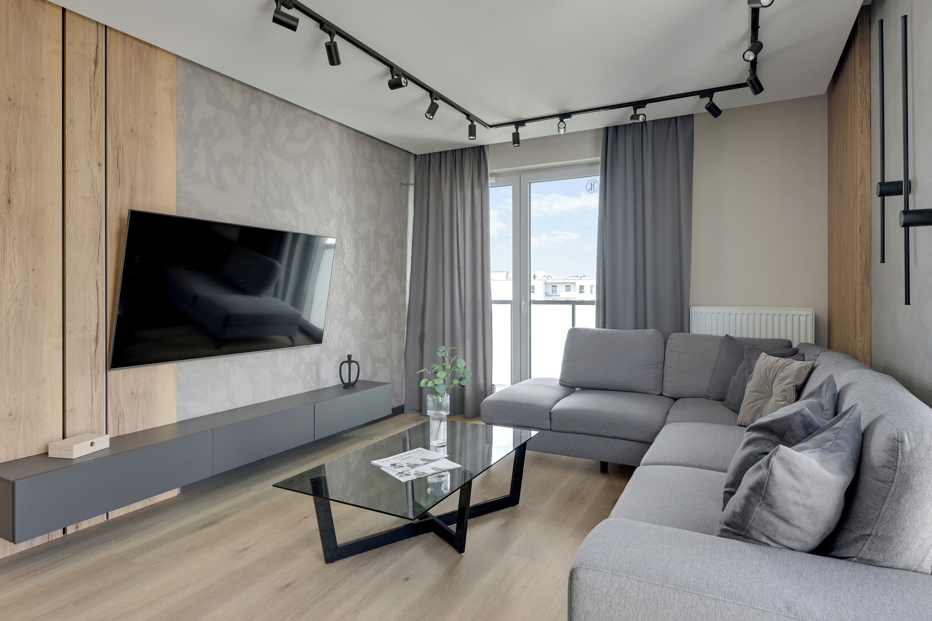 A modern living room with a large flat-screen TV mounted on a wooden panel wall, a grey sectional sofa, a glass coffee table, and a large window with curtains.