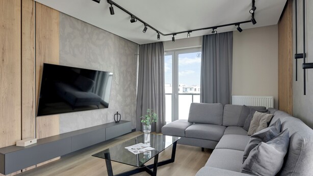 A modern living room with a large flat-screen TV mounted on a wooden panel wall, a grey sectional sofa, a glass coffee table, and a large window with curtains. - Nowa Letnica - Gdańsk Panoramic View  (Gdańsk)