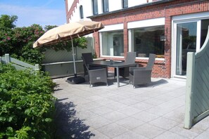 An outdoor patio with a table and chairs, a covered umbrella, and a brick building in the background. - Holiday apartment in Juist with swimming pool (Juist)