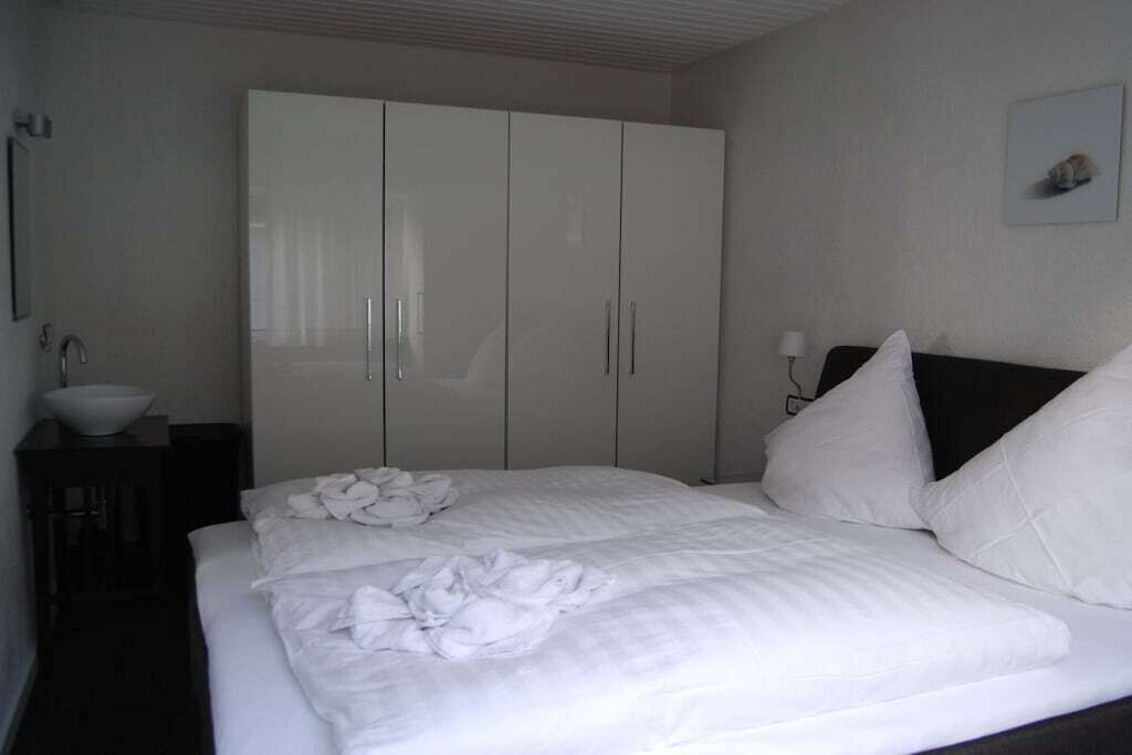 A bedroom with a bed, white linens, a large wardrobe, a bedside table with a lamp, and a framed picture on the wall.