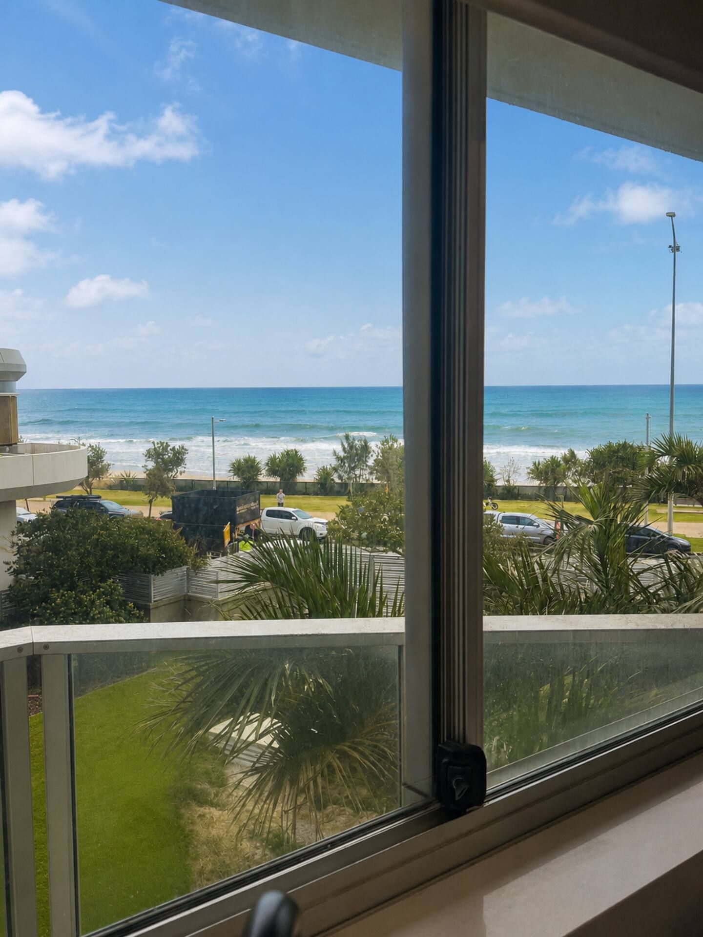 Vista dalla finestra su una spiaggia con l'oceano, auto parcheggiate e palme.