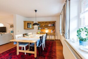 Uma área de refeições com mesa de madeira, cadeiras e tapete vermelho estampado. Há uma estante com livros e objetos decorativos, e uma janela com cortinas.