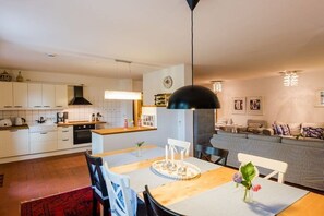 Une cuisine moderne avec des meubles blancs, un coin repas doté d’une table en bois et un espace de vie comprenant un canapé et un téléviseur.