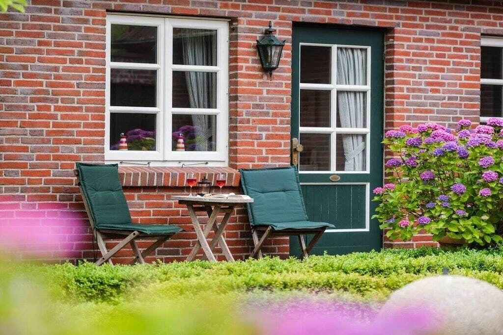 A brick building with two green chairs and a small table, a green door, and a window with curtains.