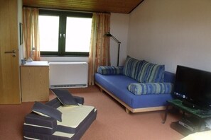 Une pièce comprenant un canapé, un téléviseur, un bureau avec une lampe et une fenêtre avec des rideaux.