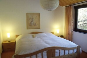 Un lit simple avec un cadre en bois et une literie blanche, dans une pièce dotée d’un plafond en bois et d’une fenêtre avec des rideaux.