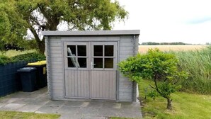 A small, grey shed with a door and windows, situated in a grassy area with a tree and a fence in the background. - Holiday home with 3 bedrooms (Neßmersiel)