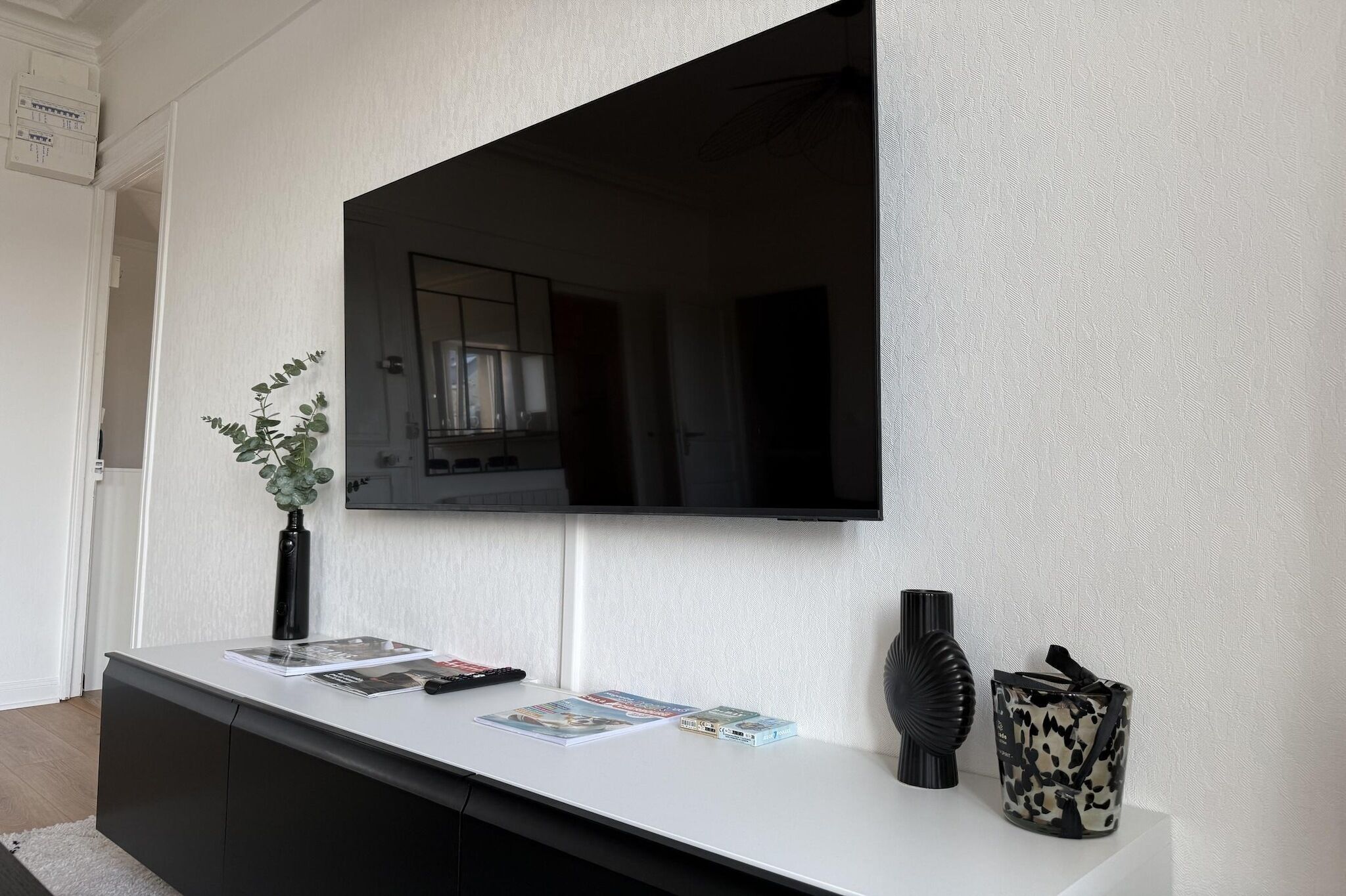Ein modernes Interieur mit einem an einer strukturierten Wand montierten Flachbildfernseher, einem weißen Konsolentisch mit Zeitschriften, einer schwarzen Vase und einem dekorativen Gegenstand.