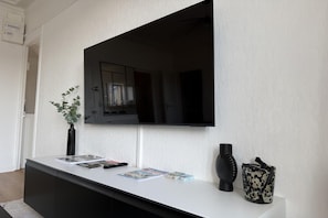 A modern interior with a flat-screen TV mounted on a textured wall, a white console table with magazines, a black vase, and a decorative item. - Nice Apartment in Cherbourg for 4 Guests (Cherbourg-en-Cotentin)