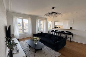 A modern living room with a dark sofa, a coffee table with a vase of flowers, and a kitchen area with bar stools. - Nice Apartment in Cherbourg for 4 Guests (Cherbourg-en-Cotentin)