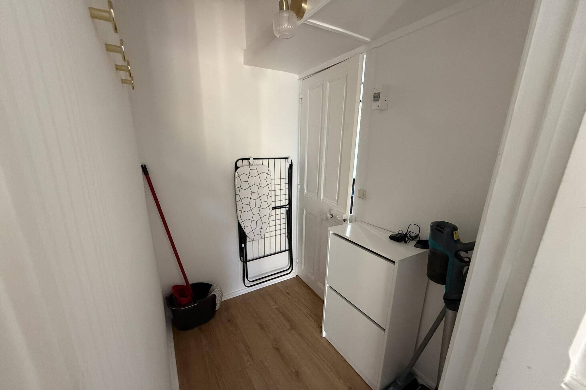 Un couloir étroit, avec un balai appuyé contre le mur, un étendoir métallique et une porte blanche.