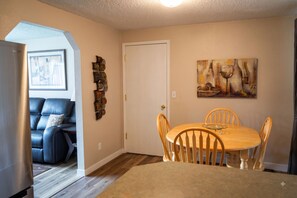 A dining area with a wooden table and chairs, a painting on the wall, and a door leading to another room.