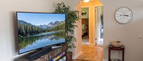 A living room with a flat-screen TV on a wooden stand, a bookshelf with board games, a potted plant, and a clock on the wall.