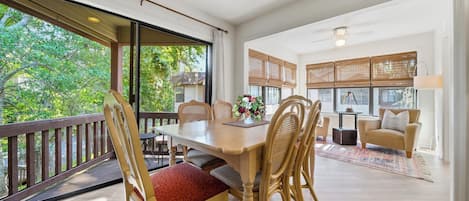 A spacious dining area with a large table and chairs, a balcony with a railing, and a view of greenery outside.