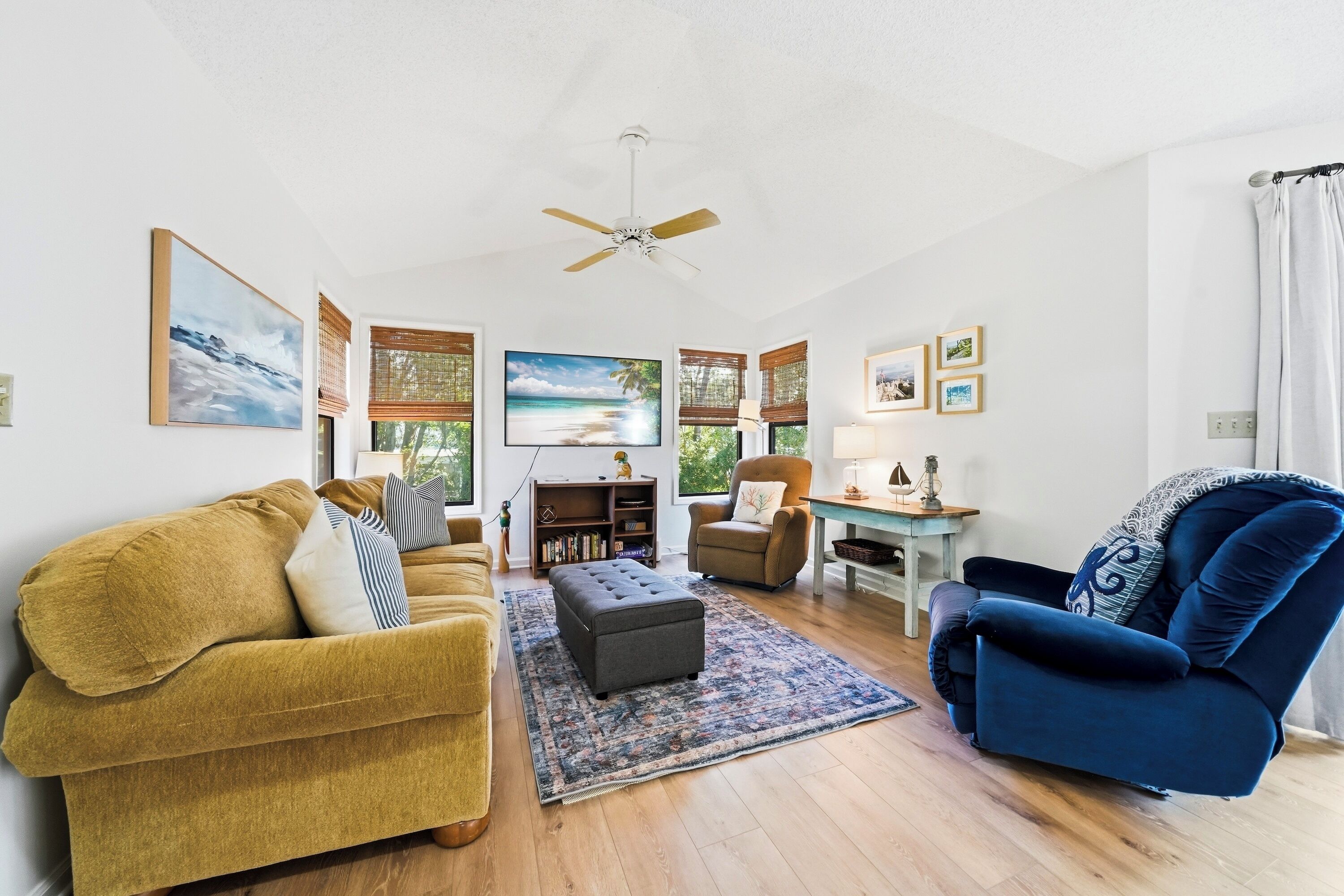 A living room with a sofa, armchair, ottoman, and a coffee table. There is a ceiling fan, a TV, and a bookshelf.
