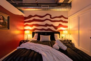A hotel room with a bed, two bedside tables, a nightstand, a lamp, and a painting on the wall. - 4BR 3BA KC Loft Collection Rooftop Bar Spa (Kansas City)