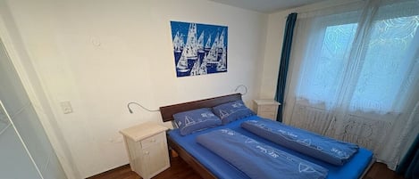 Ein Schlafzimmer mit einem Bett, einem Nachttisch, einem Wandbild und einem Fenster mit Vorhängen.