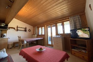 Une pièce chaleureuse avec un plafond en bois, une table à manger recouverte d’une nappe rouge, un téléviseur et une fenêtre avec des rideaux.