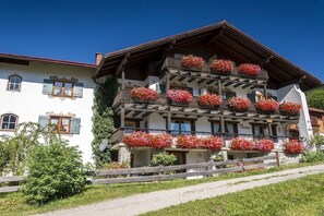 Ein mehrgeschossiges Gebäude mit Blumen geschmückten Balkonen und einem hölzernen Zaun davor, unter einem wolkenlosen Himmel.