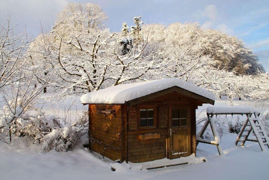A wooden cabin with a snow-covered roof, surrounded by snow-laden trees and a wooden swing set.