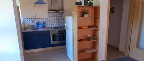 A compact kitchen with a stove, oven, and a small refrigerator. There is a wooden shelf with a vase and a bowl on it.