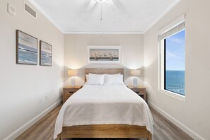 Une chambre avec un grand lit, deux tables de chevet, un ventilateur de plafond et une fenêtre donnant sur la mer.