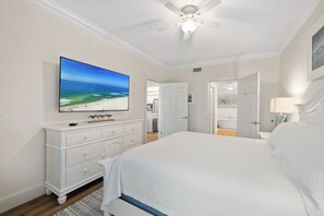Ein Schlafzimmer mit einem großen Bett, einem Deckenventilator, einem Fernseher, der eine Strandansicht zeigt, und einem durch eine offene Tür einsehbaren Badezimmer.