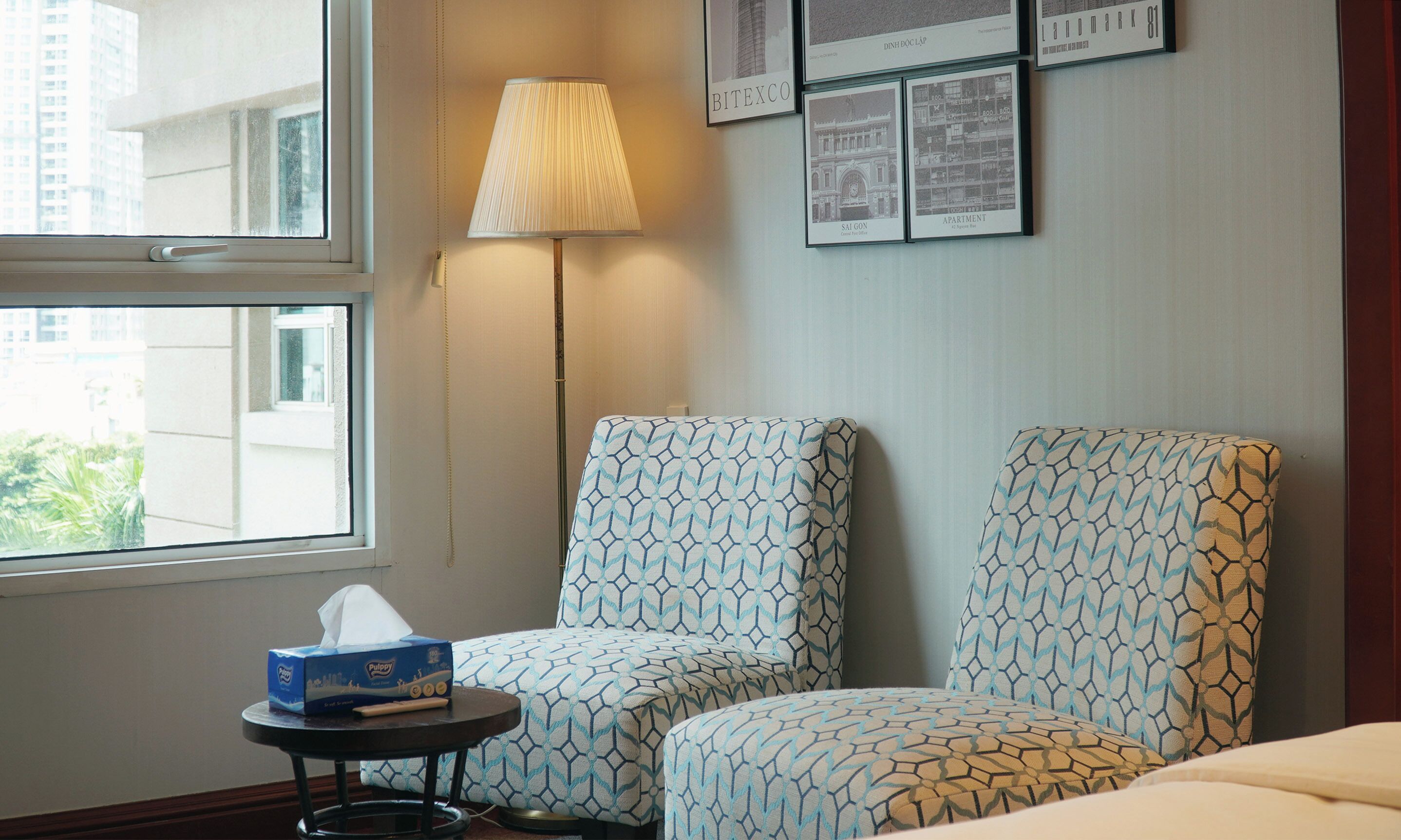 A hotel room with two patterned armchairs, a bedside table with a tissue box, a lamp, and framed pictures on the wall.