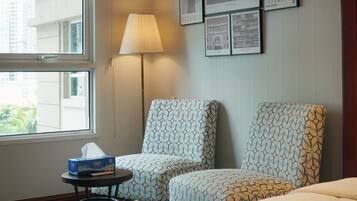 A hotel room with two patterned armchairs, a bedside table with a tissue box, a lamp, and framed pictures on the wall.