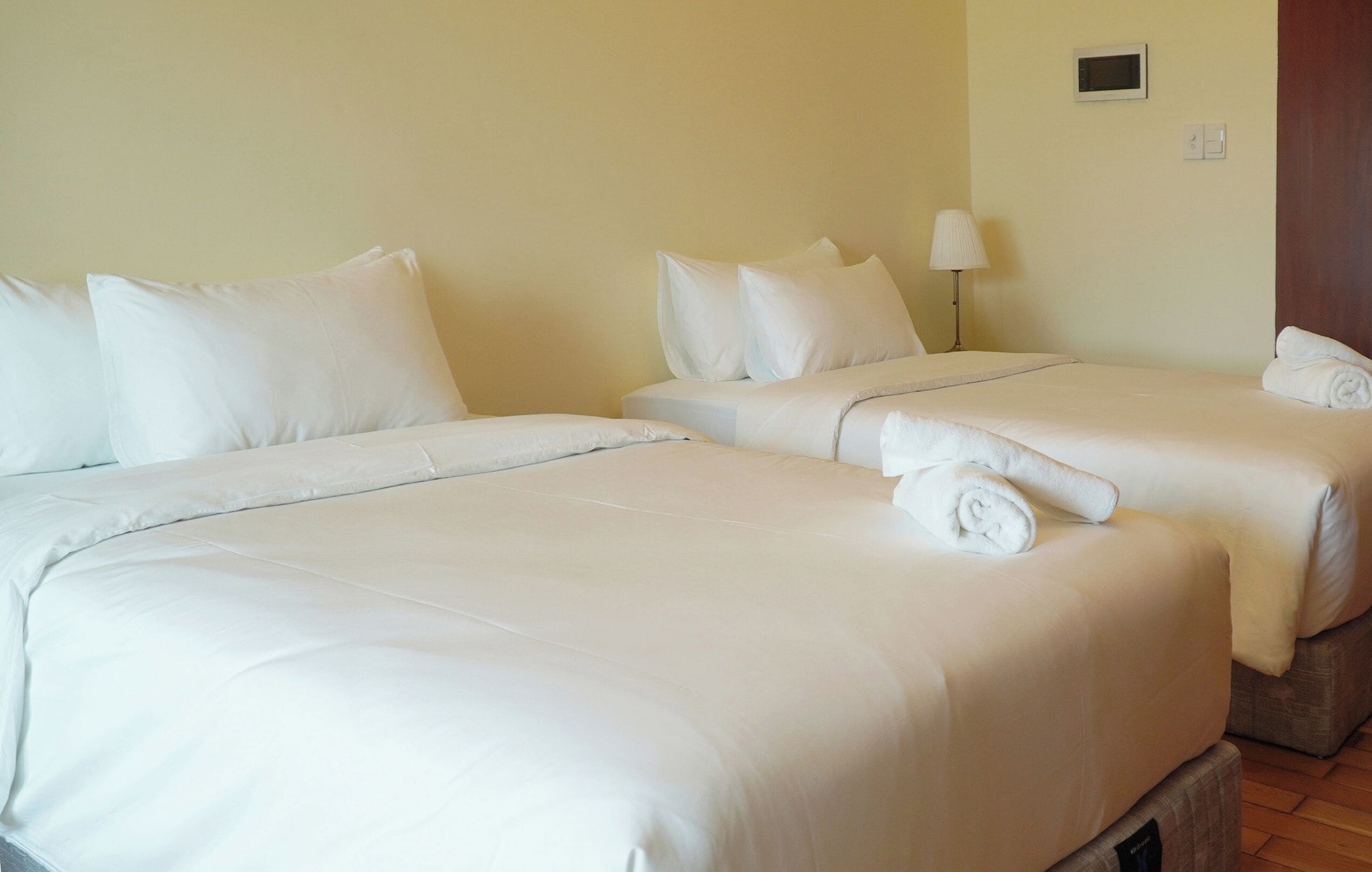 A hotel room with two beds, each with white bedding and pillows, a bedside lamp, and a rolled towel on one bed.