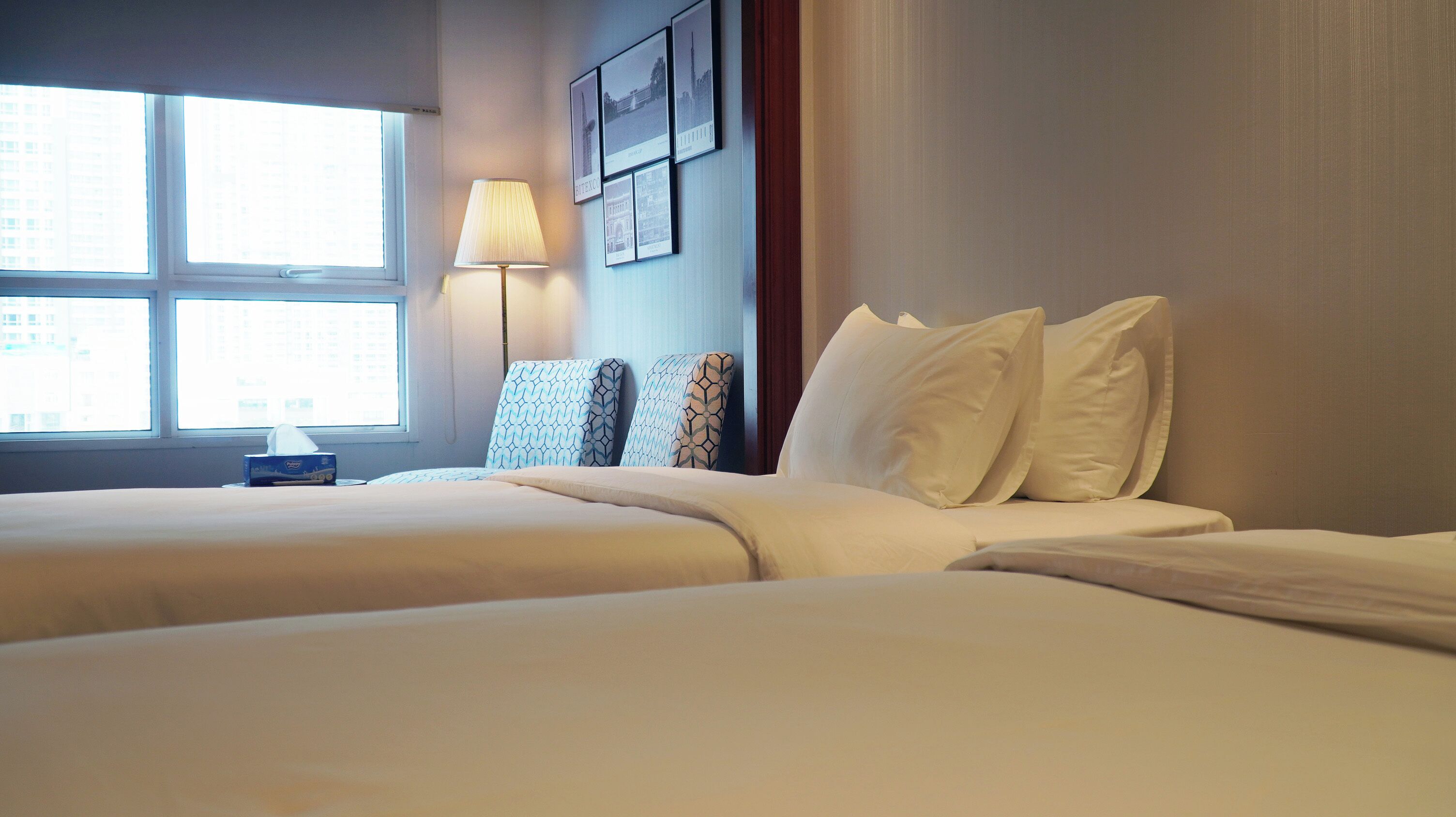 A hotel room with two beds, a bedside table, a lamp, and a window with a view of buildings.