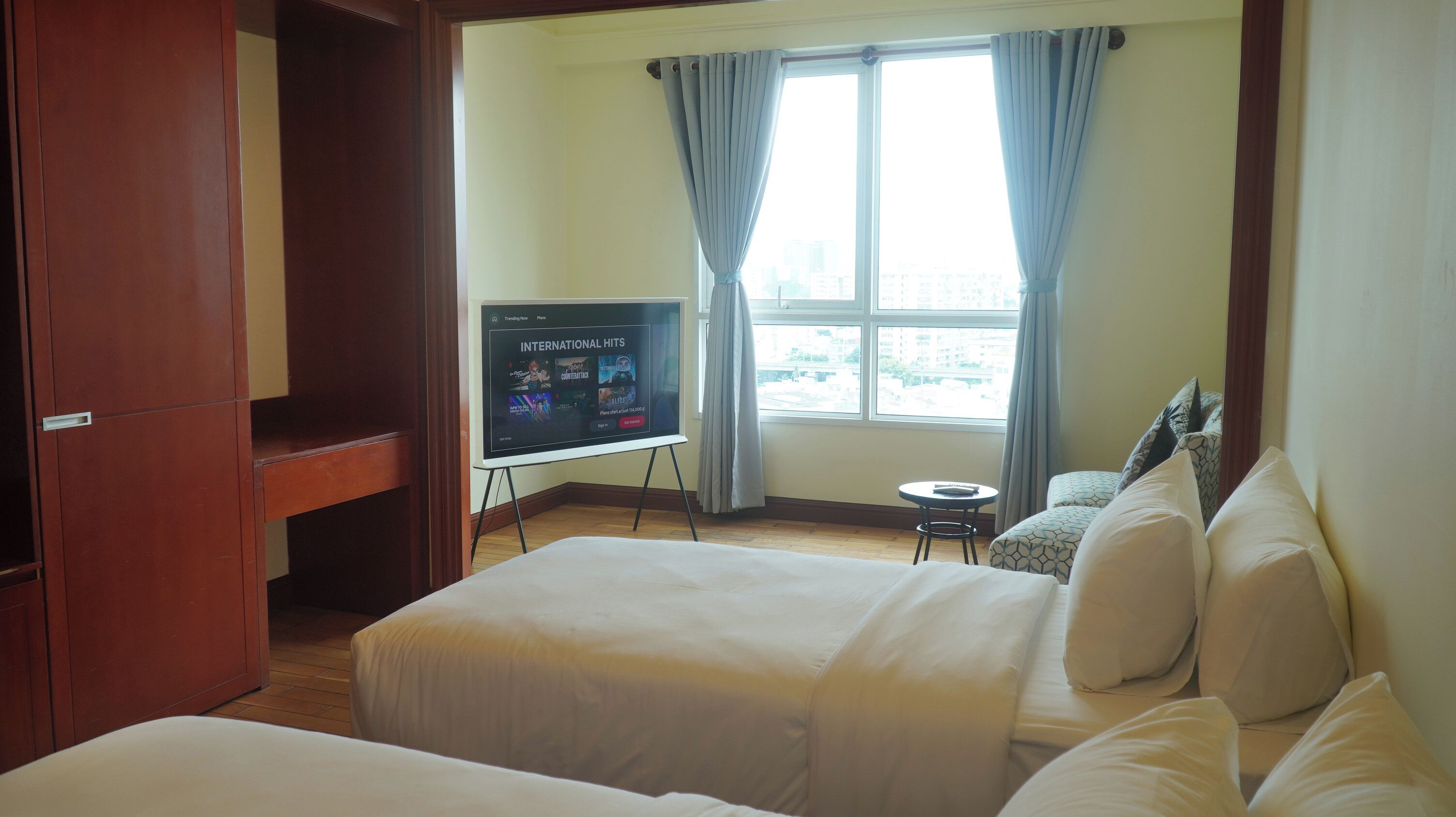 A hotel room with a bed, a television on a stand, a window with curtains, and a small table.