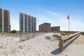 Een zandstrand met een houten loofpad, een hek en hoge gebouwen op de achtergrond.