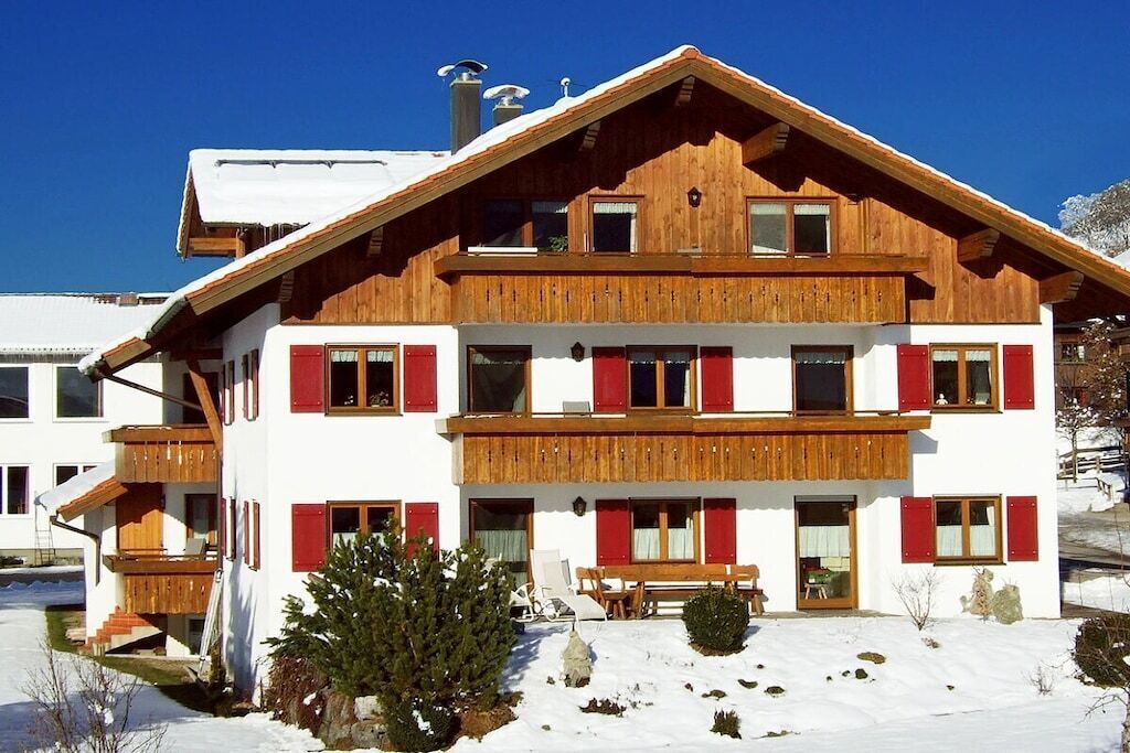 A two-story wooden chalet with red shutters, a balcony, and a snow-covered ground.