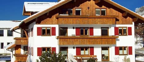 A two-story wooden chalet with red shutters, a balcony, and a snow-covered ground.