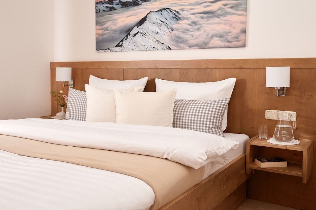 Ein Hotelzimmer mit einem hölzernen Kopfteil, weißer Bettwäsche, einem karierten Kopfkissen, einem Nachttisch mit Glas und Krug sowie einer Wandlampe.