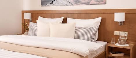 A hotel room with a wooden headboard, white bedding, a checkered pillow, a bedside table with a glass and a pitcher, and a wall-mounted lamp.