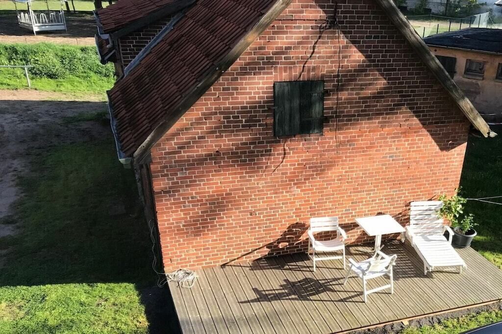 Um edifício de tijolos com telhado inclinado, uma pequena área de deck com mobiliário de exterior branco e um vaso de planta.