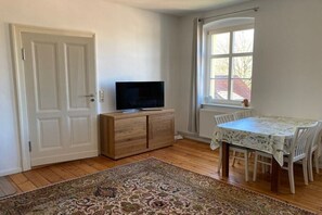 Ein Zimmer mit Fernseher auf einer Holzkommode, einem Esstisch mit Stühlen, einem gemusterten Teppich und einem Fenster mit Vorhängen.