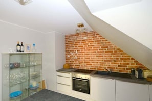 A compact kitchen with a brick wall, a built-in oven, and a glass-fronted shelving unit. - Knutt in the house Bill15 (Juist)