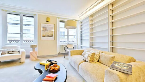 A modern living room with a sofa, a coffee table with books and a bowl of fruit, a chair, and a large window with blinds. - AN EXCELLENT COMBINATION OF PARISIAN CHARM & CENTRAL CONVENIENCE - SAINT GERMAIN (Paris)