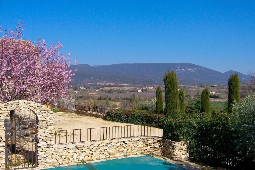 Une piscine en pierre, bordée d’un muret en pierre et protégée par une clôture métallique, le tout entouré de verdure et offrant une vue sur des montagnes lointaines.