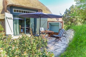 A patio with a table and chairs, a striped chair, and a thatched-roof building in the background. - Friesenhaus Sara (S4) (Wenningstedt)
