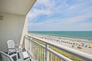 Ein Balkon mit weißen Stühlen und einem Tisch, der auf einen Strand mit Sonnenschirmen und Menschen blickt.