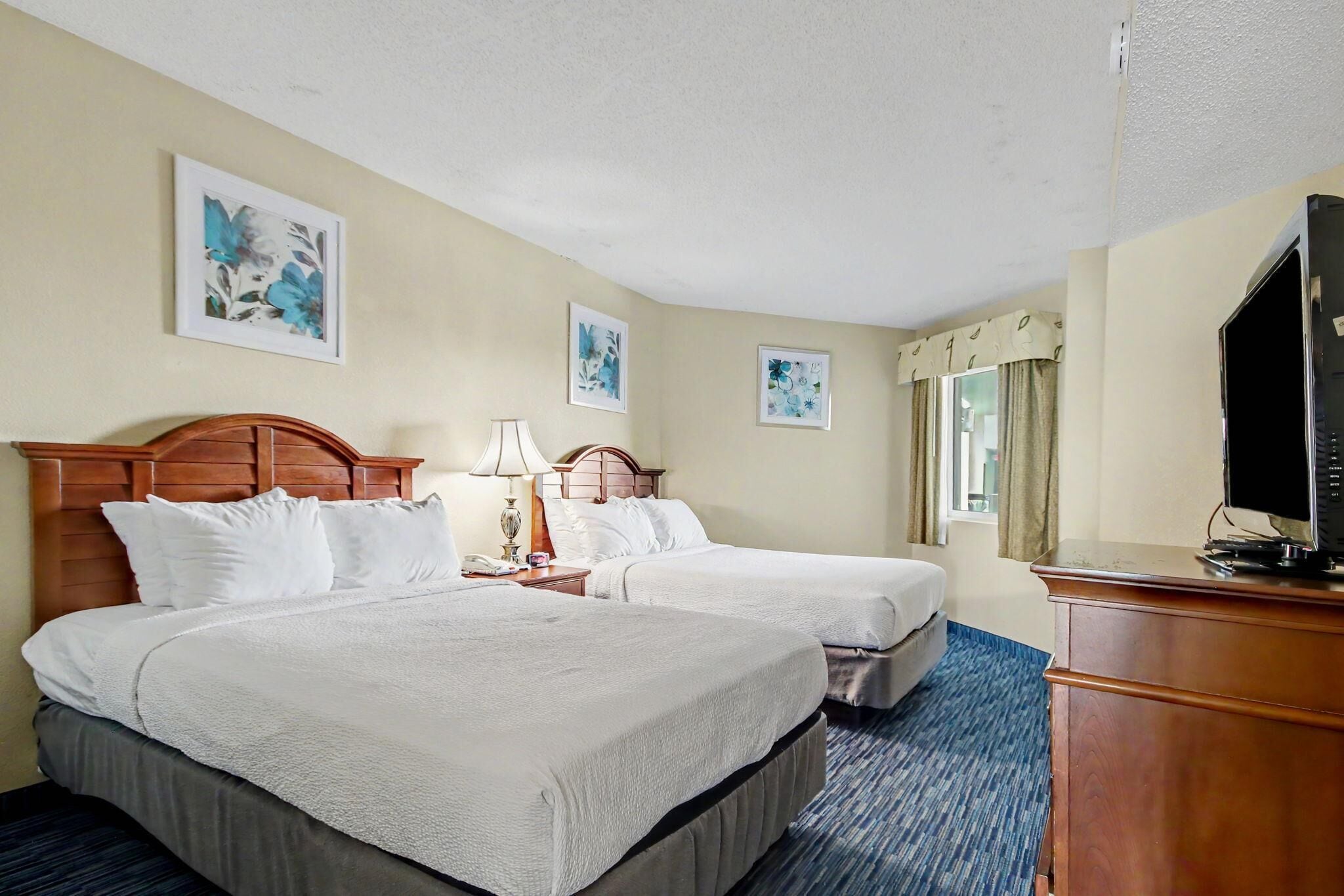 A hotel room with two beds, a desk, a television, and artwork on the walls.