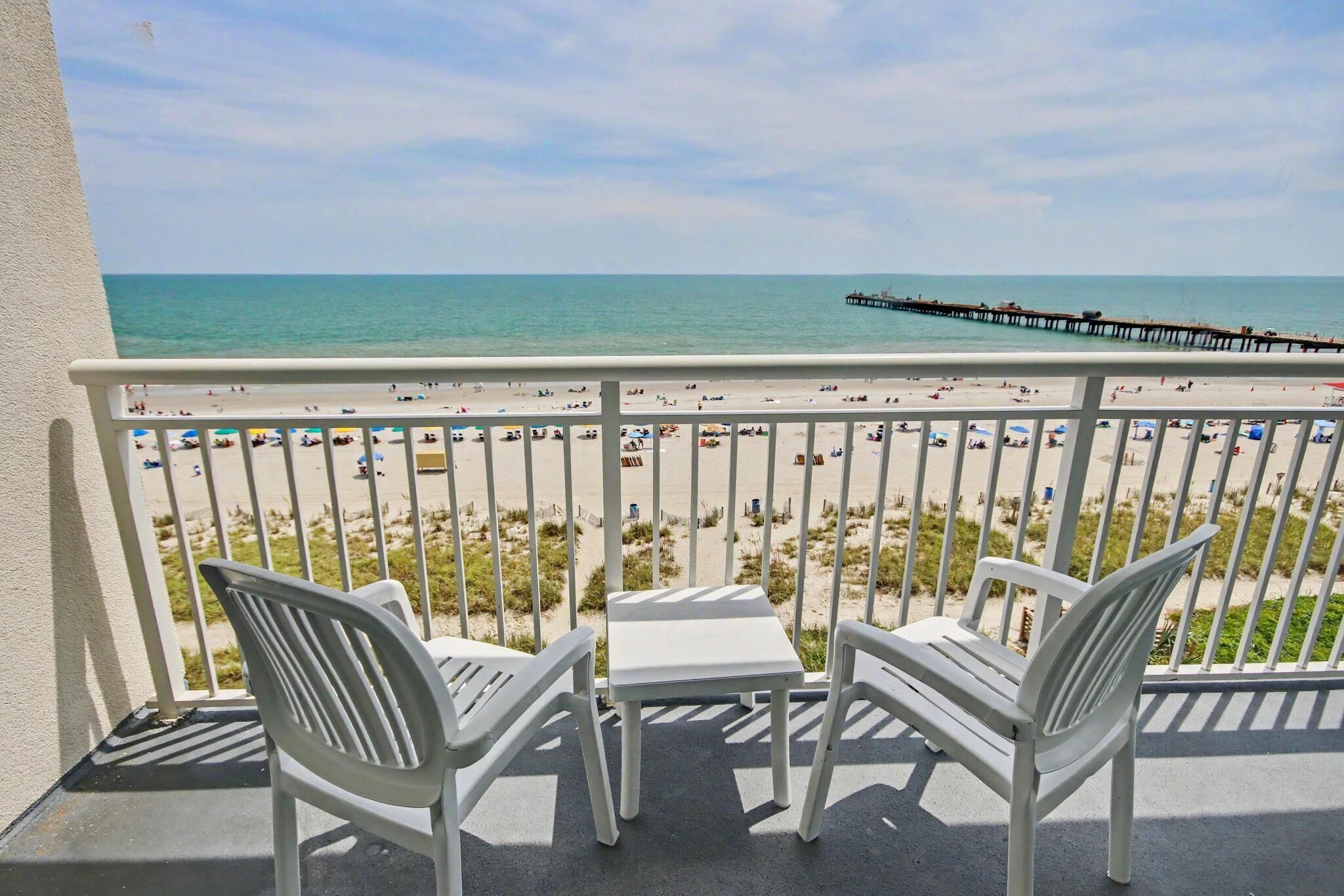 A balcony with two white chairs and a small table, overlooking a beach with a pier in the distance.