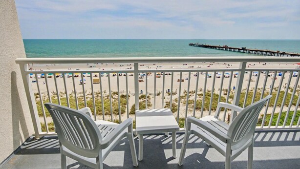 Ein Balkon mit zwei weißen Stühlen und einem kleinen Tisch, mit Blick auf einen Strand und einen Pier im Hintergrund.